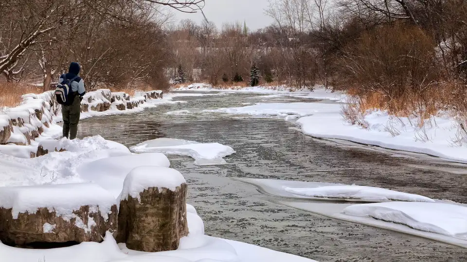 Person mit Rucksack am verschneiten Ufer eines Flusses im Winter